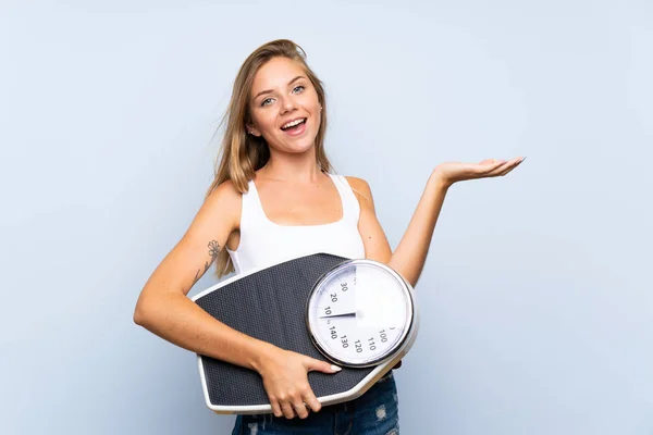 Young blonde girl with weighing machine over isolated blue white background — ストック写真