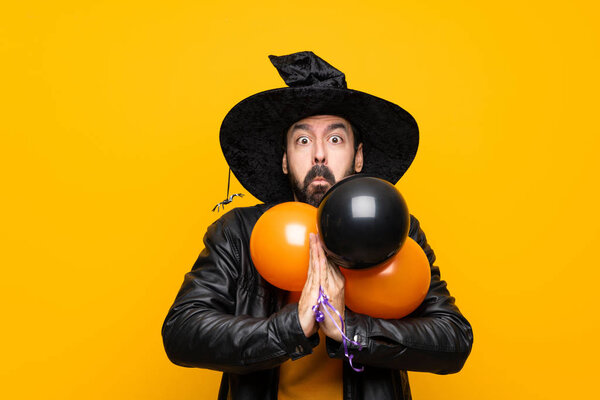 Man with witch hat holding black and orange air balloons for halloween party keeps palm together. Person asks for something