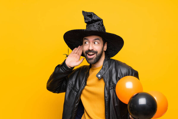 Man with witch hat holding black and orange air balloons for halloween party listening to something by putting hand on the ear