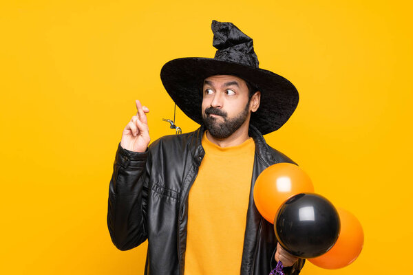 Man with witch hat holding black and orange air balloons for halloween party with fingers crossing and wishing the best