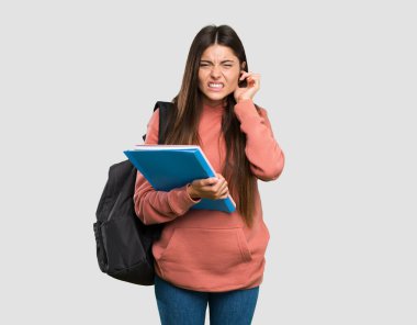 Elinde defterler tutan genç bir öğrenci kız hayal kırıklığına uğramış ve gri arka planda kulaklarını kapatmış.