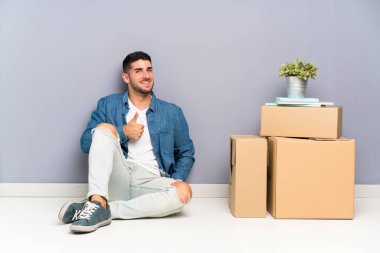 Yakışıklı genç adam kutular arasında yeni bir evde hareket jest bir başparmak veren