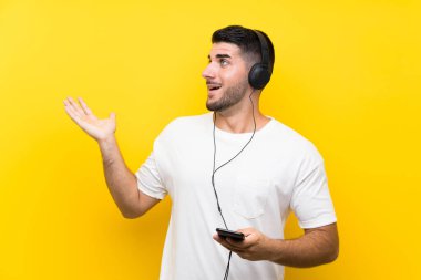 Genç yakışıklı adam sürpriz yüz ifadesi ile izole sarı duvar üzerinde bir cep ile müzik dinliyor