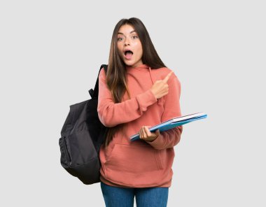 Elinde not defterleri olan genç bir bayan gri arka plana şaşırmış ve onu işaret ediyor.