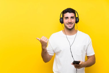 Genç yakışıklı adam izole sarı duvar üzerinde bir cep ile müzik dinleme yan bir ürün sunmak için işaret