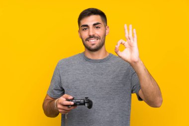 Genç yakışıklı bir adam video oyunu kontrolörüyle oynuyor. İzole edilmiş sarı arkaplan üzerinde. Parmaklarıyla tamam işareti gösteriyor.