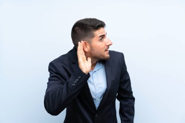 Mavi arka planda yakışıklı bir adam kulağına elini sürerek bir şeyler dinliyor.