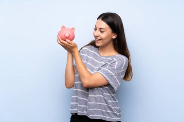 Büyük bir kumbara tutan izole mavi arka plan üzerinde genç esmer kadın