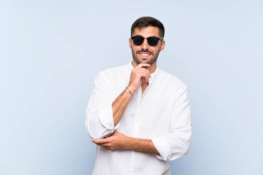 Gözlüklü ve gülümseyen, soyutlanmış mavi arka planda sakallı yakışıklı bir adam.