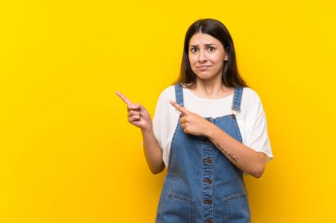 İzole edilmiş sarı arka planı olan genç bir kadın. Korkmuş ve yan tarafı işaret ediyor.