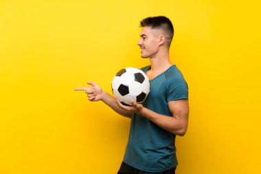 Yakışıklı genç futbolcu izole sarı arka plan üzerinde yan bir ürün sunmak için işaret