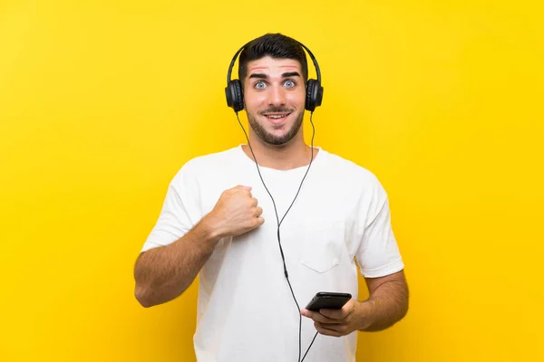 驚きの表情で孤立した黄色の壁の上にモバイルで音楽を聴く若いハンサムな男 — ストック写真