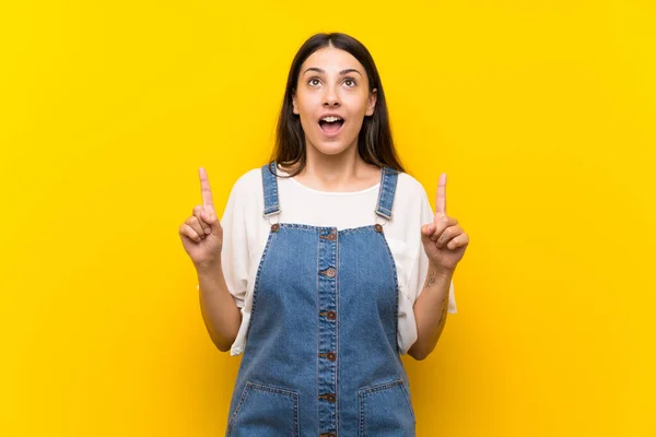 Jovem Mulher Dungarees Sobre Isolado Amarelo Fundo Surpreso Apontando Para — Fotografia de Stock