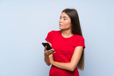 Mavi duvarda cep telefonu olan esmer bir kadın gülümserken yukarı bakıyor.