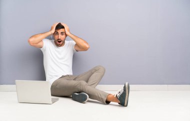 Dizüstü bilgisayarı olan genç bir adam şaşırmış bir yüz ifadesiyle yerde oturuyor.