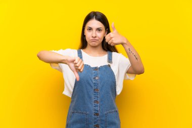 İzole edilmiş sarı arka plan üzerinde kot giyen genç bir kadın iyiye-kötüye işaret yapıyor. Evet ya da hayır arasında kararsız