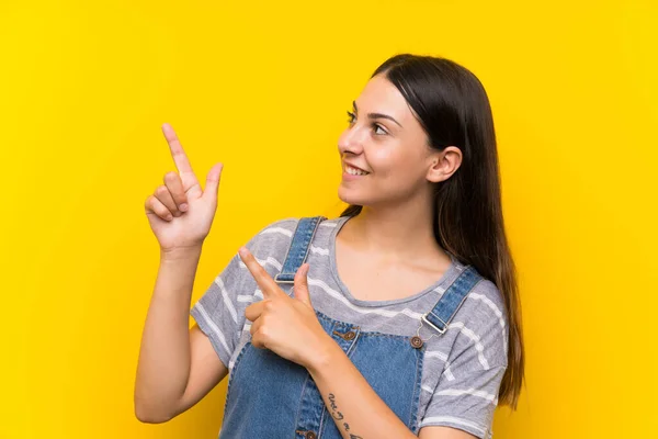 Jovem Mulher Dungarees Sobre Fundo Amarelo Isolado Apontando Com Dedo — Fotografia de Stock