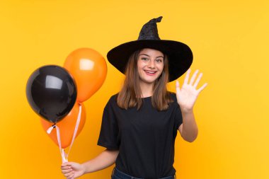 Cadılar bayramı partileri için elinde siyah ve turuncu hava balonları tutan genç cadı mutlu bir ifadeyle selamlıyor.