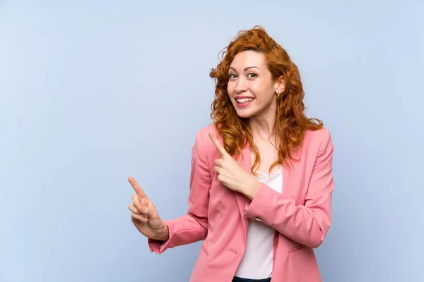 Redhead woman in suit over isolated blue wall frightened and pointing to the side