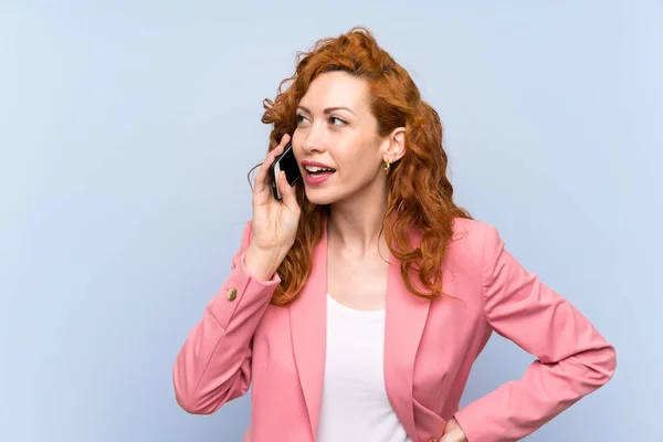 Redhead woman in suit over isolated blue wall keeping a conversation with the mobile phone