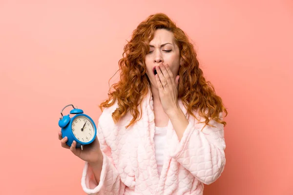 Redhead woman in dressing gown holding vintage clock and yawning — Stock Photo, Image