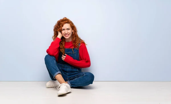 Redhead woman with overalls sitting on the floor having doubts