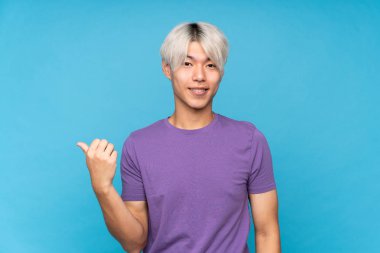 İzole edilmiş mavi arka planda genç Asyalı bir adam bir ürünü sunmak için kenarı işaret ediyor.