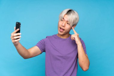 İzole edilmiş mavi arka planda genç Asyalı bir adam selfie çekiyor.