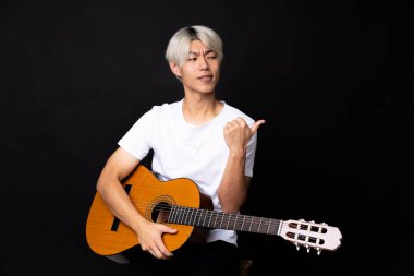 Siyah arka planda gitarı olan genç Asyalı bir adam bir ürünü sunmak için kenarı işaret ediyor.