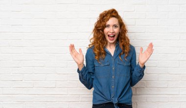 Suratı şoke olmuş beyaz tuğlalı duvarın üzerinde kızıl saçlı bir kadın.