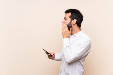 Sakallı genç adam cep telefonu tutuyor ve ağzı sonuna kadar açık bağırıyor.