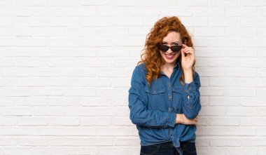 Kızıl saçlı kadın beyaz tuğlalı duvarın üzerinde gözlüklü ve mutlu