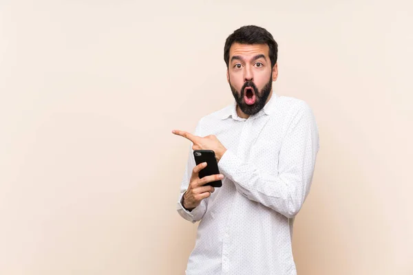 Joven Con Barba Sosteniendo Móvil Sorprendido Señalando Lado —  Fotos de Stock