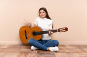 Mladá žena s kytarou sedící na podlaze s pochybnostmi a zmateným výrazem ve tváři
