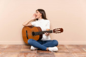 Mladá žena s kytarou sedí na podlaze a křičí s ústy dokořán