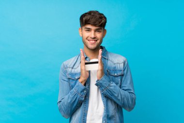 Soyutlanmış mavi arka planı olan genç yakışıklı bir adam elinde kredi kartı tutuyor.