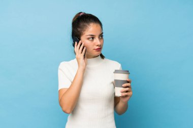 Genç esmer kız izole edilmiş mavi arka planda elinde kahve ve bir cep telefonu var.