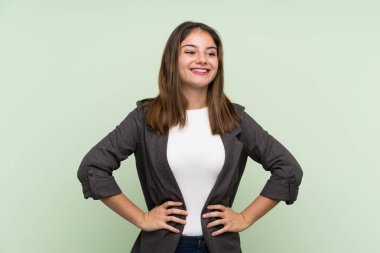 Siyah ceketli genç esmer kız izole edilmiş yeşil arka planda kolları kalçasında ve gülümsüyor.