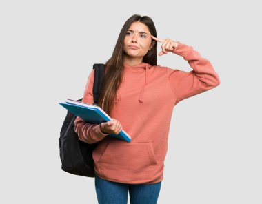 Elinde not defterleri olan genç bir kız deliliğin jestini yapıyor. Gri arkaplanı parmağıyla kapatıyor.