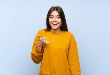 Kafkasyalı genç bir kadın izole edilmiş mavi bir duvarda telefon jesti yapıyor.