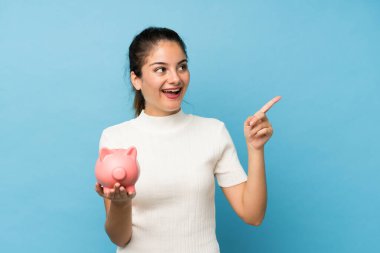 Genç esmer kız izole edilmiş mavi arka planda büyük bir kumbara tutuyor.