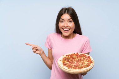 İzole edilmiş mavi duvarın üzerinde pizza tutan genç ve güzel bir kız şaşırmış ve parmağıyla kenarı işaret ediyor.