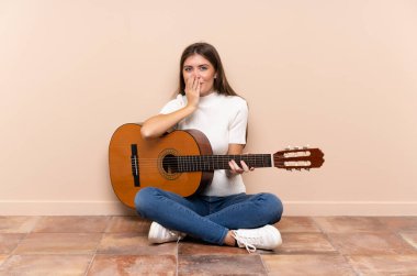 Gitarlı genç bir kadın yerde oturuyor ve yüzünde sürpriz bir ifade var.
