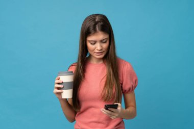 İzole edilmiş mavi arka planda, elinde kahve ve cep telefonu olan genç bir kadın.