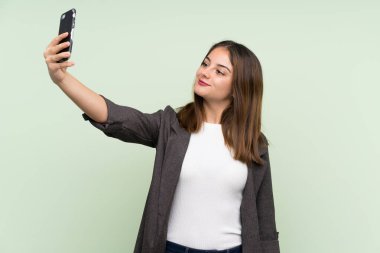 Yeşil arka planda ceketi olan esmer genç bir kız selfie çekiyor.
