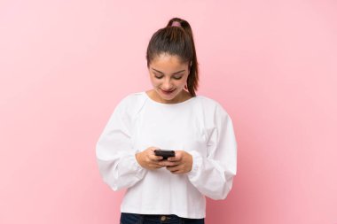 İzole edilmiş pembe arka planda genç esmer kız cep telefonuyla bir mesaj gönderiyor.