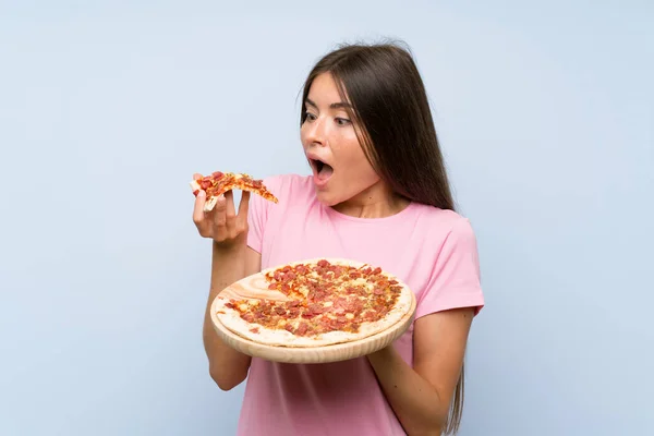 Красивая молодая девушка держит пиццу над изолированной голубой стеной — стоковое фото