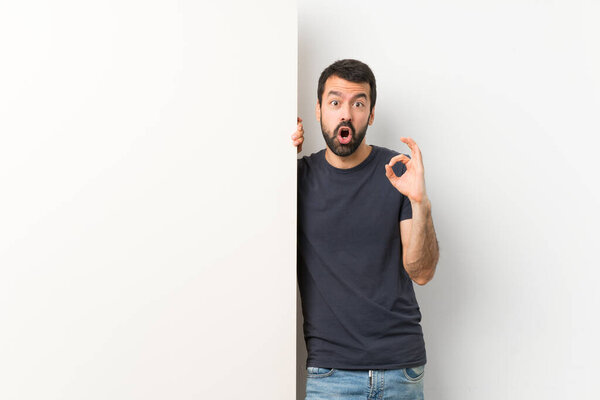 Young handsome man with beard holding a big empty placard surprised and showing ok sign