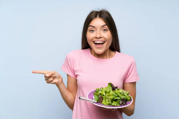 Jonge Vrouw Met Salade Geïsoleerde Blauwe Achtergrond Verrast Wijzend Vinger — Stockfoto