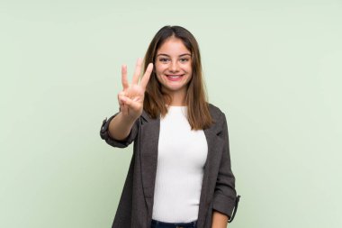 Yeşil arka planda ceketi olan esmer genç kız mutlu ve parmaklarıyla üçe kadar sayıyor.